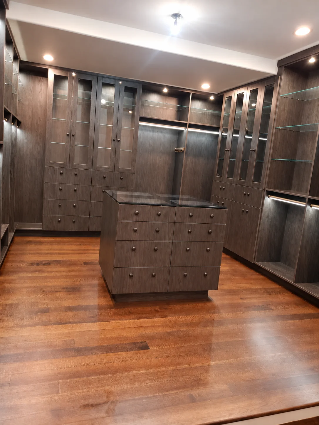 Dark wood-grain dressing room with glass cabinet doors, glass shelves, lighting and a glass-top drawer island