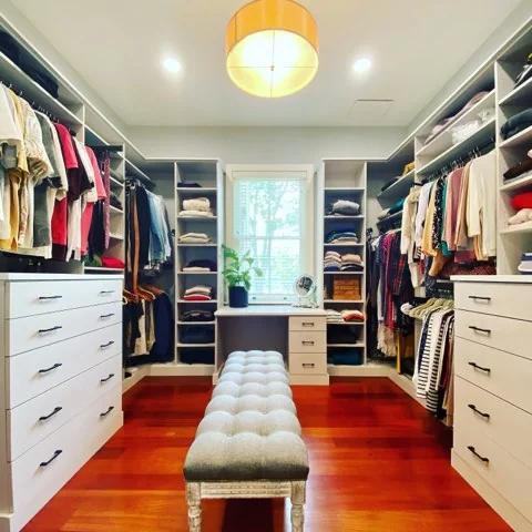 Walk-in closet in use with pale drawer cabinets, hanging clothes, open shelving, a dressing table beneath the window and a tufted bench