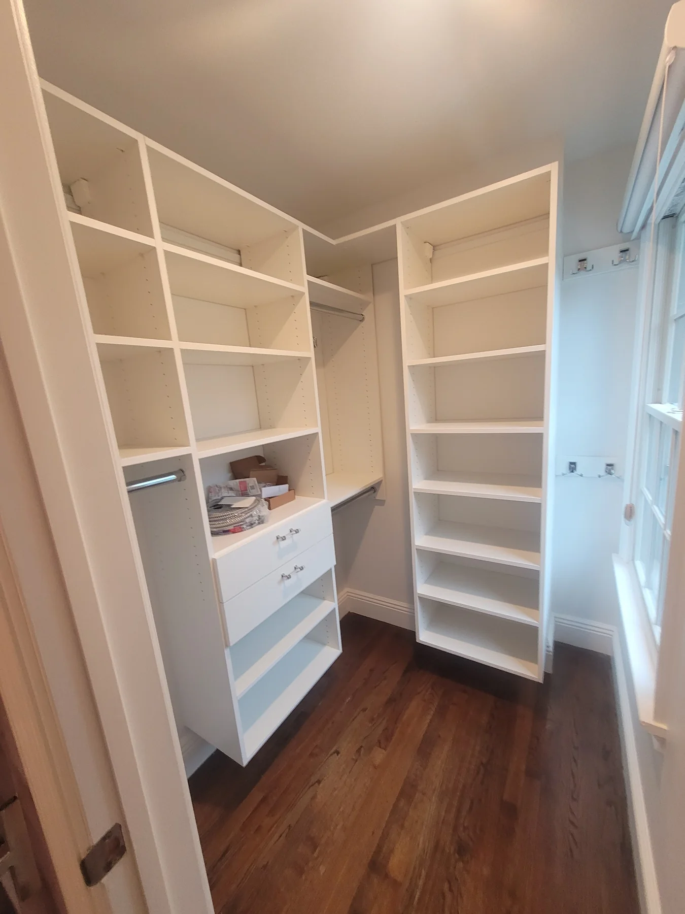 During installation: white corner closet with hanging rails, two drawers and adjustable shelves beside a window