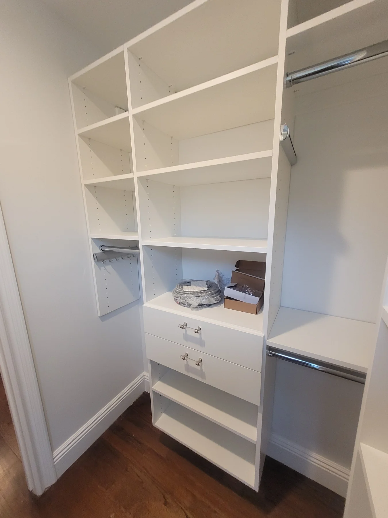 During installation: white closet drawer cabinet with clear handles, shelves and hanging storage while finishing supplies remain on the work surface