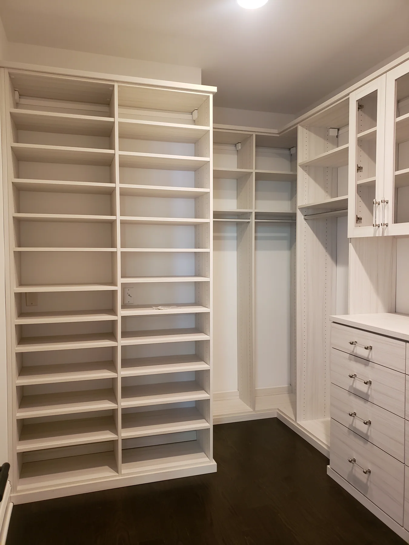 White walk-in closet with a tall shoe tower, wood-accented cabinetry and dark flooring